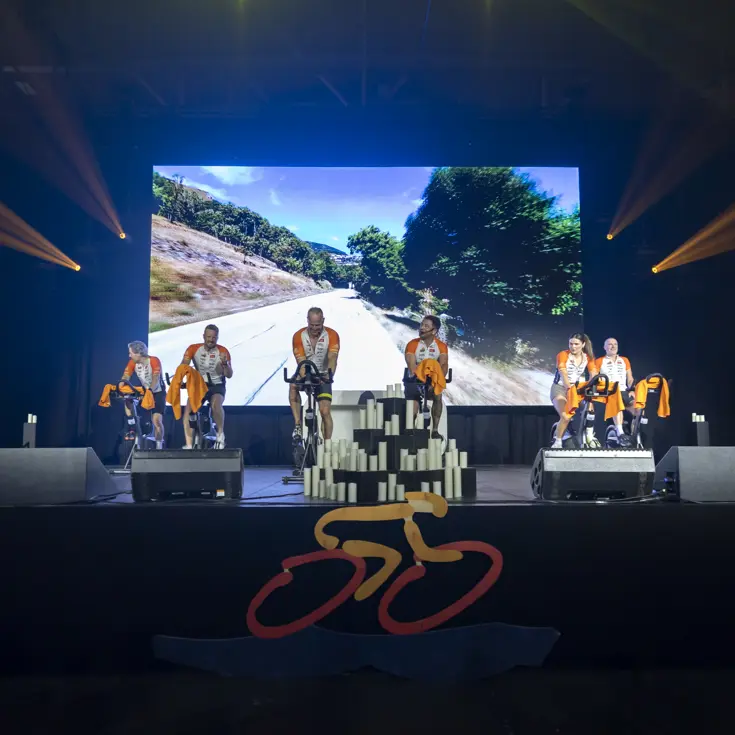 Deelnemers van team Marechaussee aan de Alpe d’HuZes Indoorride 2026 op een podium met beeldscherm erachter. 