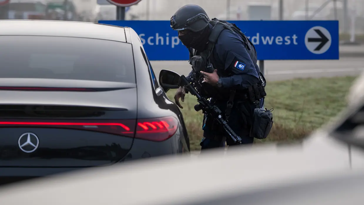 Een bewapende marechaussee in donker uniform buigt bij een zwarte Mercedes, met een blauw bord "Schiphol Noordwest" en een snelheidsbeperkingsbord op de achtergrond.