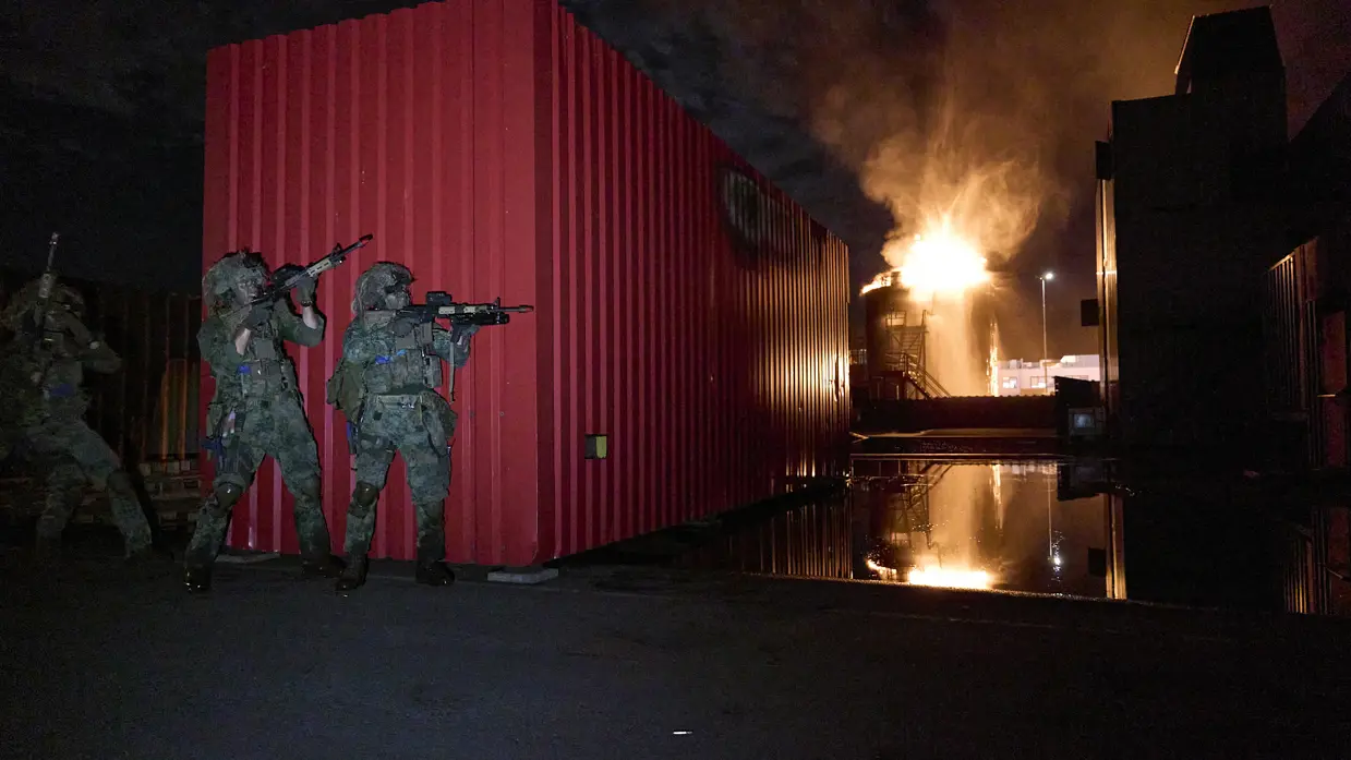 3 mariniers schuilen achter een container terwijl er om de hoek brand lijkt te woeden op een verhoogde installatie.