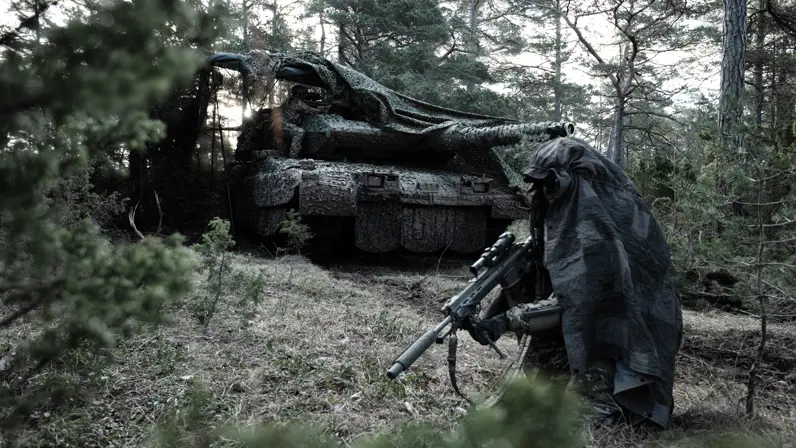 Een soldaat en een gepantserd militair voertuig in het bos, beiden gecamoufleerd.