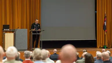 Alttekst: Links: een groep mannen luistert naar een man in uniform op een podium. Rechts: 2 mannen in uniform kijken naar een munt in hun handpalm met drie man om zich heen. 