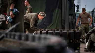 Soldaten in uniform werken met zware metalen onderdelen bij een groene militaire vrachtwagen, omringd door gereedschap en uitrusting.