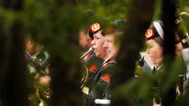 Een groene, wazige achtergrond met muzikanten in donkergroene uniformen en rode baretten die instrumenten bespelen. 