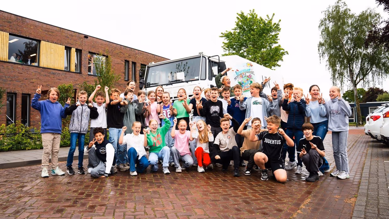 +alttekst: Groep blije kinderen en volwassenen poseren voor een witte bus met kleurrijke tekeningen, bij een rood bakstenen gebouw. 