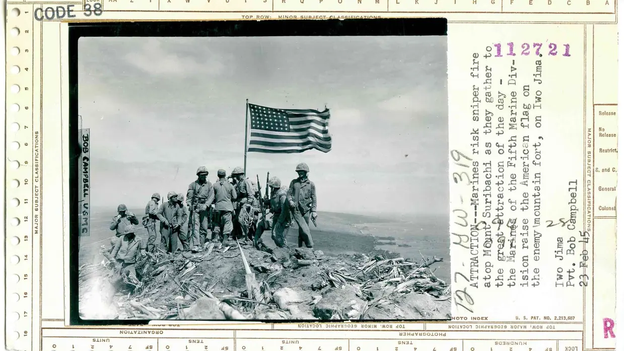 Zes Amerikaanse soldaten hijsen samen de Amerikaanse vlag op een heuveltop tijdens een veldslag. De lucht is bewolkt, de omgeving is rotsachtig en ruig.