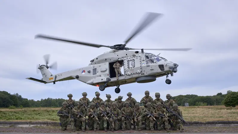 Een groep mariniers poseert voor een NH90 die achter hen in de lucht hangt.