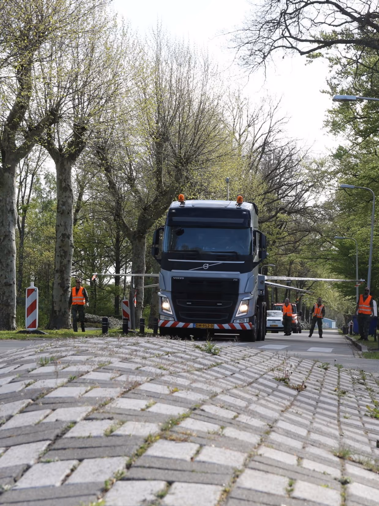 Een grote vrachtwagen rijdt over een weg met tegels, omringd door bomen en verkeersborden, met arbeiders in oranje vesten. 