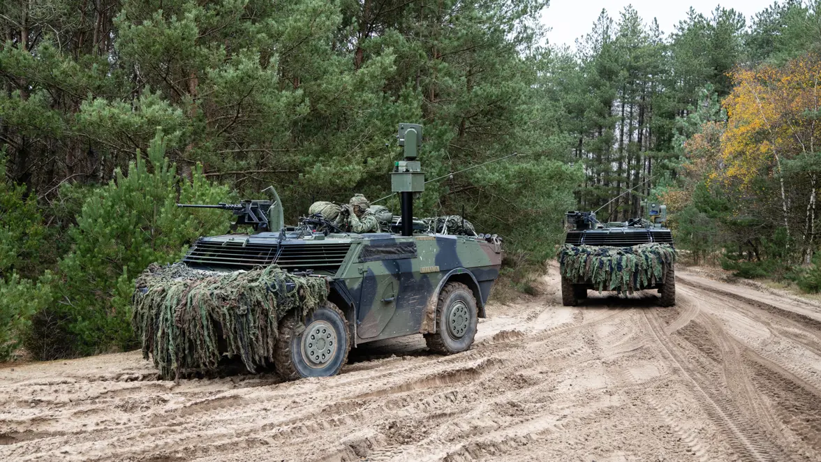 Twee militaire pantservoertuigen, bedekt met camouflagenetten, rijden over een zanderig bospad, omringd door dichte groene bomen en struiken. 