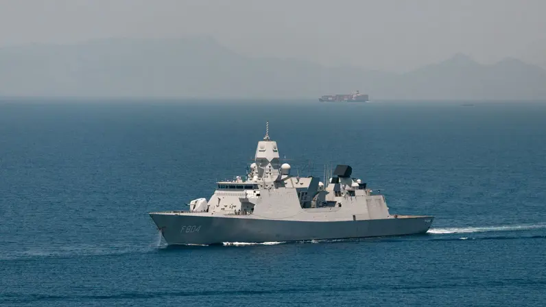 Een marineschip vaart op zee met in de verte een ander schip en hoge bergen.