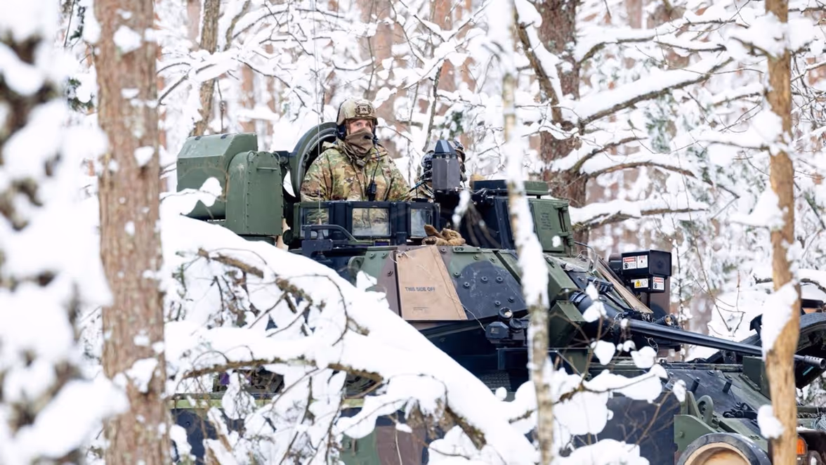 Een soldaat in winterkleding zit bovenop een gepantserd militair voertuig tussen besneeuwde bomen, omringd door sneeuw en rustige, witte natuur. 