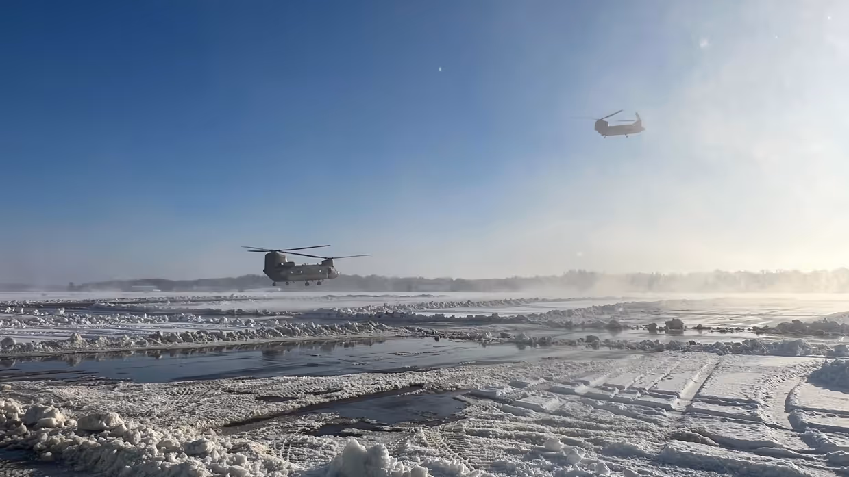 Twee helikopters vliegen laag over een besneeuwde, modderige en ijsbedekte landingsbaan onder een heldere, blauwe hemel met zonnig licht. 