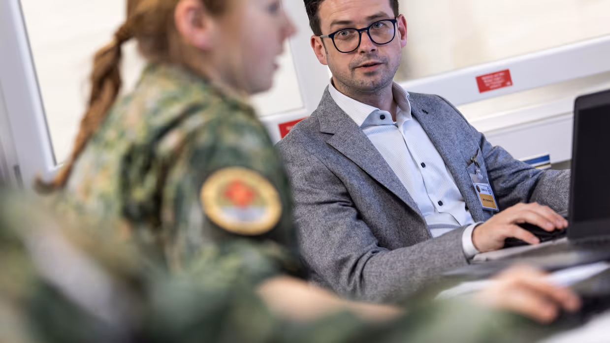 Links: Een man in pak met bril en identiteitskaart werkt op een laptop. Een vrouw in militair uniform met een medisch embleem staat naast hem. Rechts: drie mannen lopen glimlachend door een gang: een in donkerblauw uniform, een in Nederlands militair tenue met medisch embleem, en een in Amerikaans legeruniform. 