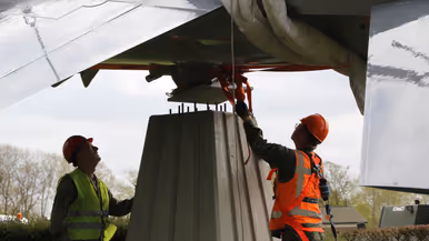 Twee arbeiders in veiligheidskleding bevestigen een groot metalen voorwerp onder de vleugel van een vliegtuig. 
