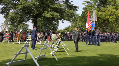 Een Amerikaanse militair staat in de houding tegenover een Nederlandse. In de achtergrond staan kransen en publiek. Rechts staan vier opgestelde militairen met vlaggen en zit het orkest van de luchtmacht.