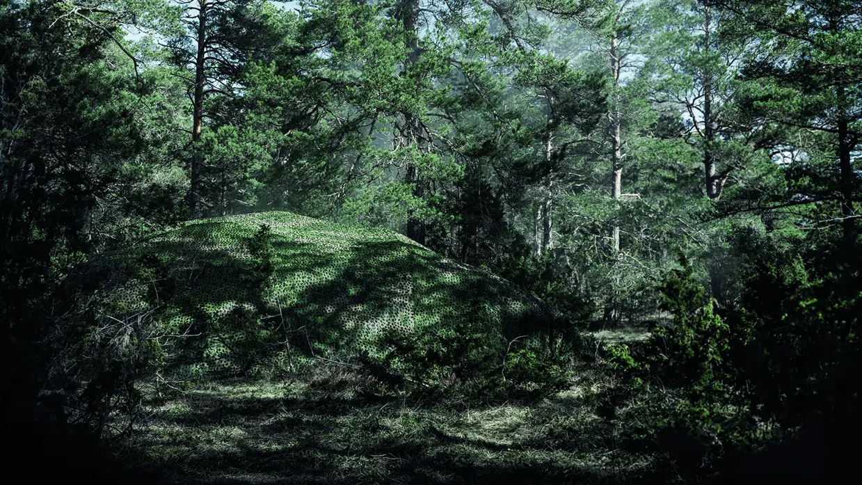 Een militair voertuig in het bos verstopt onder een groot camouflagenet.