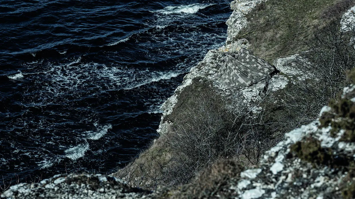 Enkele militairen verstopt op een berg aan zee onder een camouflagedeken.