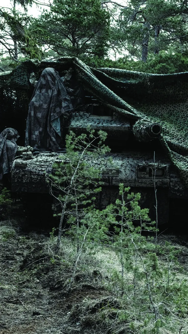 Militair voertuig in bos onder camouflagenet met daaromheen een aantal gecamoufleerde militairen.