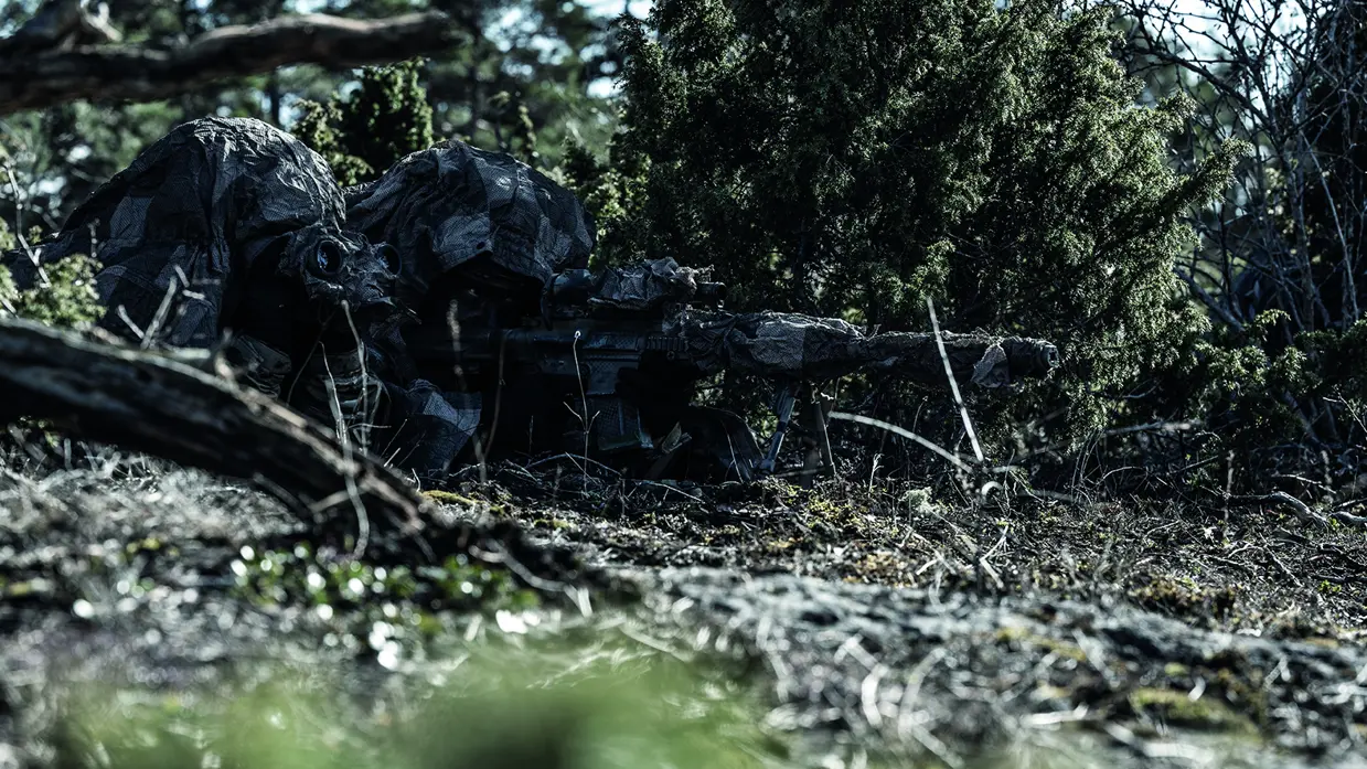 Een gecamoufleerde militair ligt achter zijn wapen in het bos.