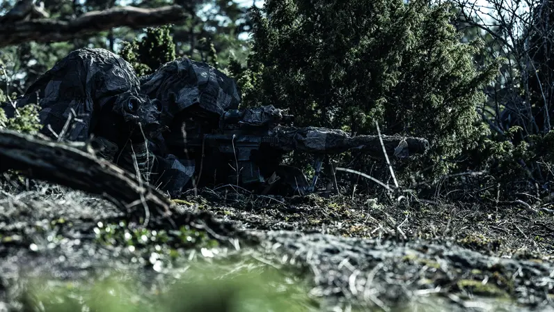 Een gecamoufleerde militair ligt achter zijn wapen in het bos.