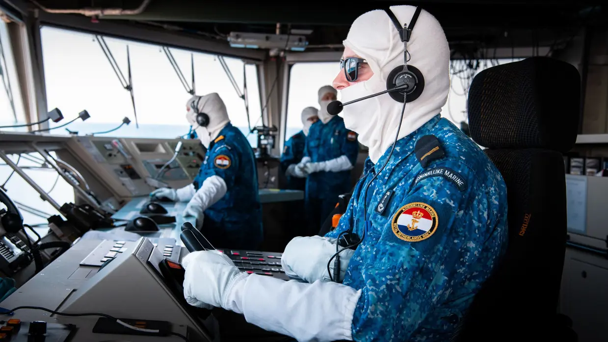 Een marineman zit op de brug van de De Ruyter en draagt antiflash-handschoenen en een antiflash-kap over zijn hoofd.