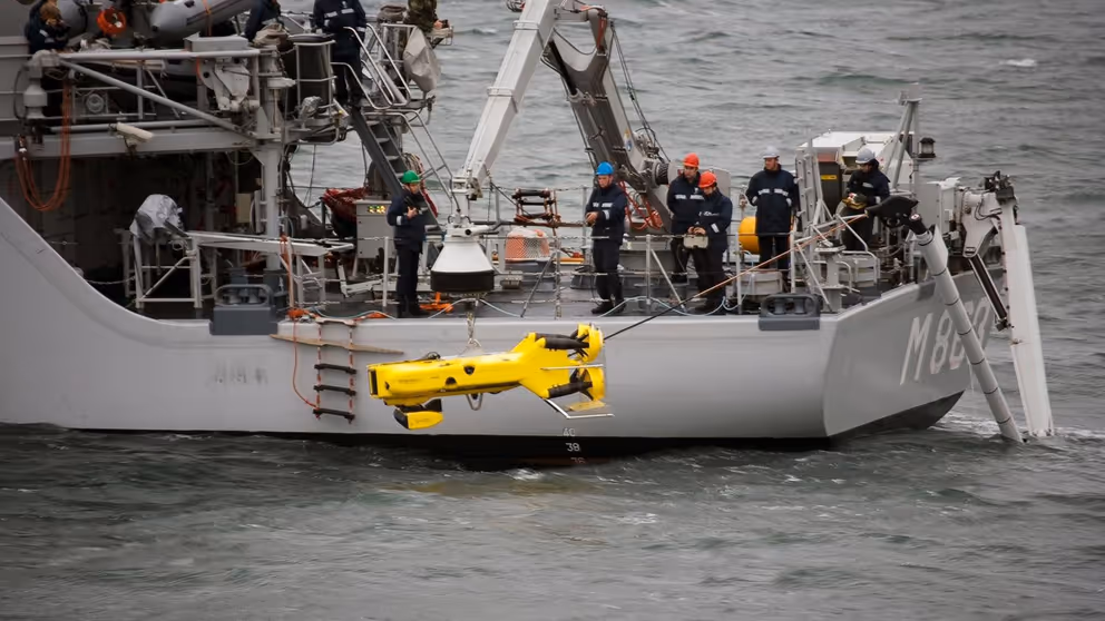 Marineschip waarop militairen een gele onderwaterrobot te water laten.