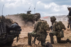 2 militairen in volledige gevechtsuitrusting beiden op een knie tussen rook. Voor hen 2 militaire voertuigen.