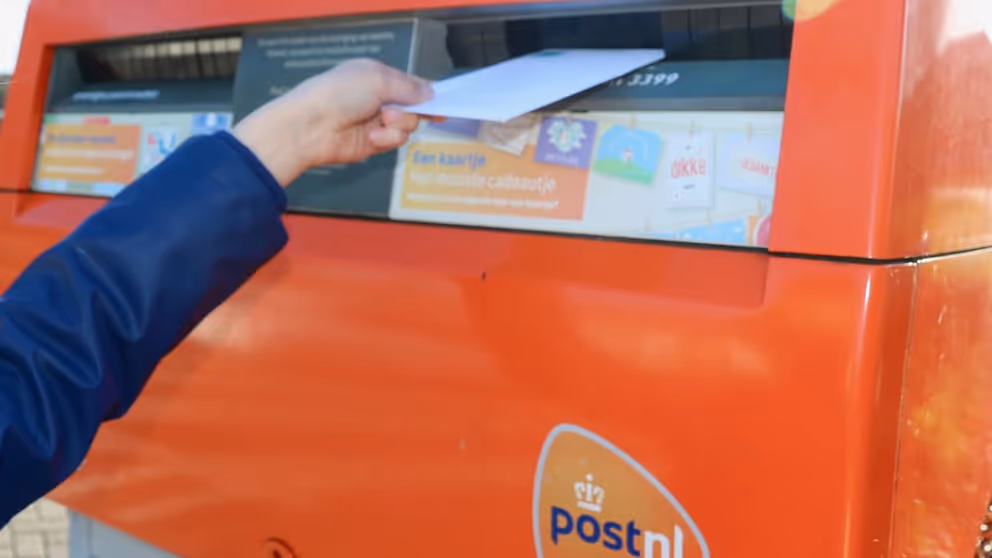 Hand gooit witte enveloppe in rode brievenbus.