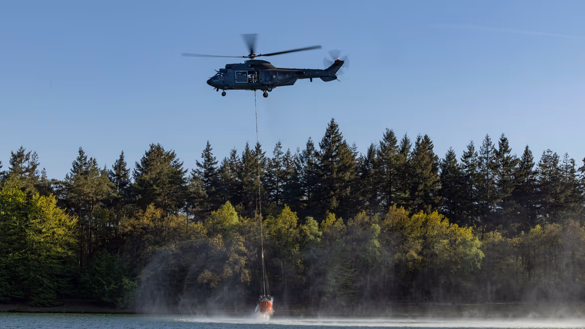 Een helikopter vliegt laag boven een meer en vult een oranje waterzak die onder de heli hangt. 
