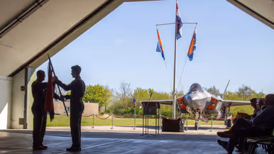 Scherders neemt de squadronvlag in ontvangst uit handen van Van de Graaf. Rechts van hen zit publiek. Op de achtergrond, net buiten de geopende shelter, staat een F-35 en een mast met drie vlaggen.