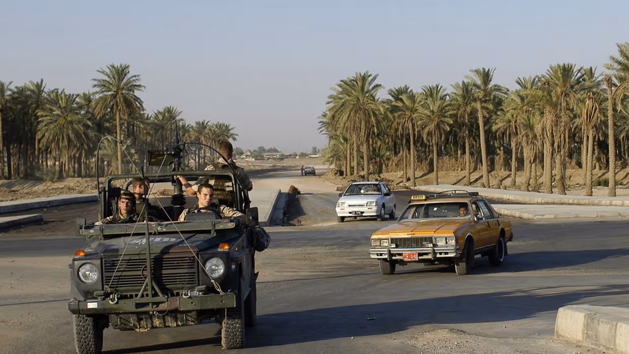 Een groene militaire jeep met soldaten rijdt op een brede weg.  Een gele taxi en een witte auto rijden mee. Palmbomen en een hoge mast op de achtergrond.