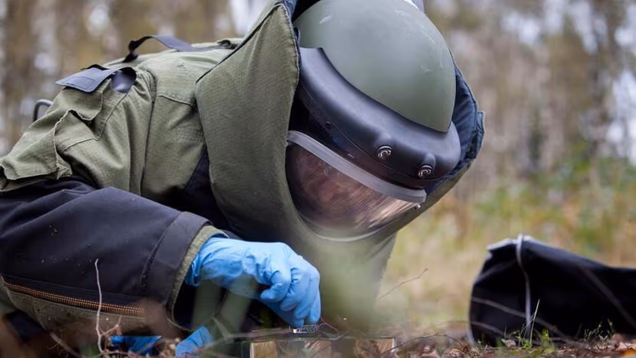 Persoon in bommenpak ligt op de grond in bos en maakt explosieven onschadelijk.