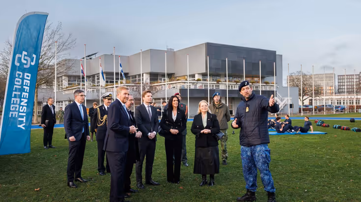 De Koning en minister staan met een aantal andere mensen bij de vlag van Defensity College.