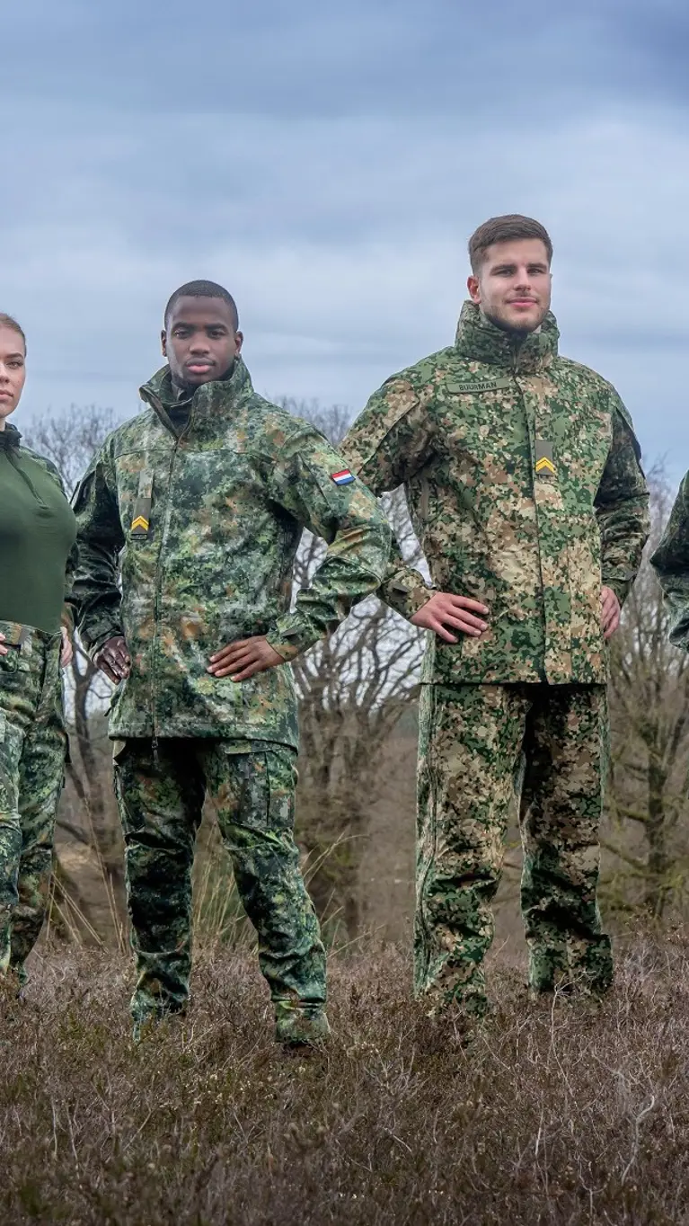 Militairen staan in het veld en dragen een vlekkenpak. Achter hen hangt een helikopter in de lucht.