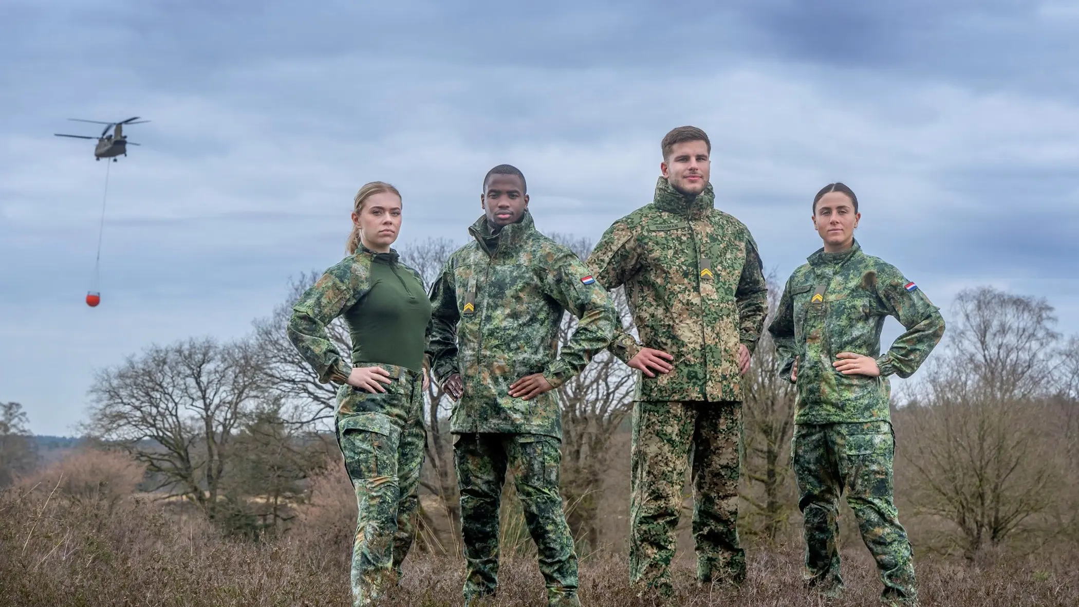 Militairen staan in het veld en dragen een vlekkenpak. Achter hen hangt een helikopter in de lucht.