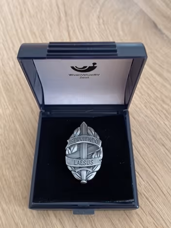 Voorbeeld van een MOD-insigne in een doosje. Het insigne bestaat uit een zwaard van mat witmetaal, omgeven door een banderol met de Latijnse tekst Serviendo Laesus.