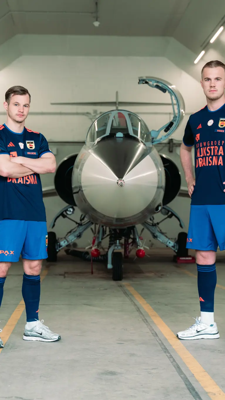 Twee mannen staan in een voetbaltenue naast een oude straaljager in een zaal.