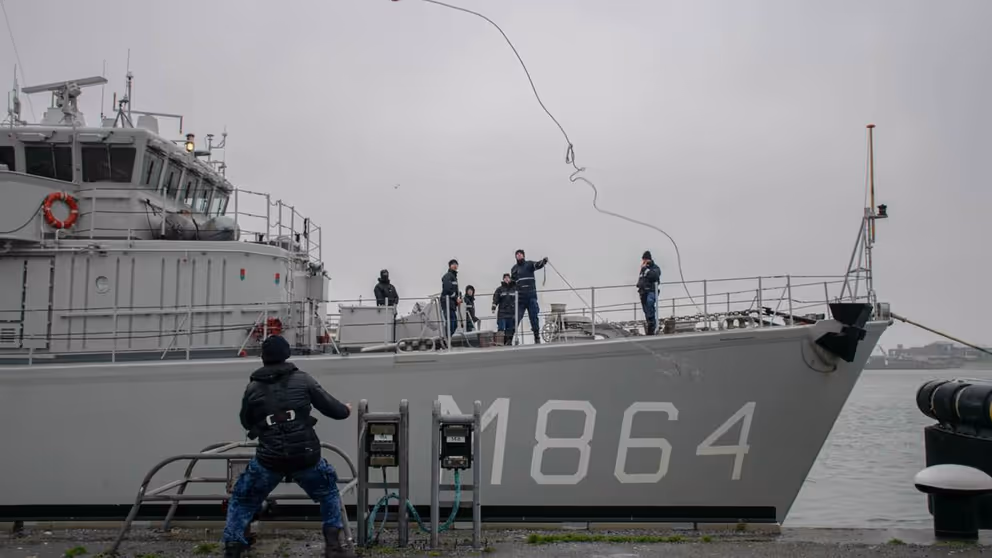 Marineschip legt aan bij kade. Militair gooit touw naar collega op kade.