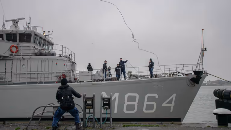Marineschip legt aan bij kade. Militair gooit touw naar collega op kade.