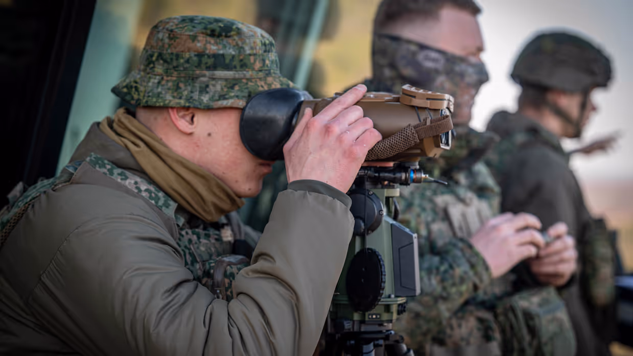Drie militairen houden overzicht op de werkzaamheden. De man op voorgrond kijkt door een stel verrekijkers.