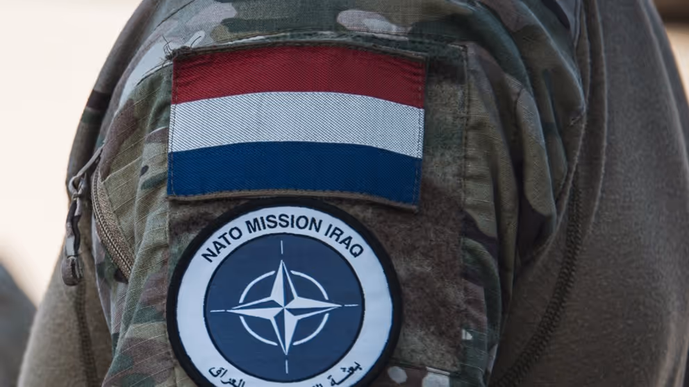 Schouder militair met NAVO-embleem en Nederlandse vlag erboven.