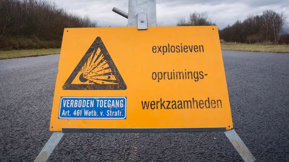 Geel bord op de weg met de tekst: explosieven opruimingswerkzaamheden en  verboden toegang.