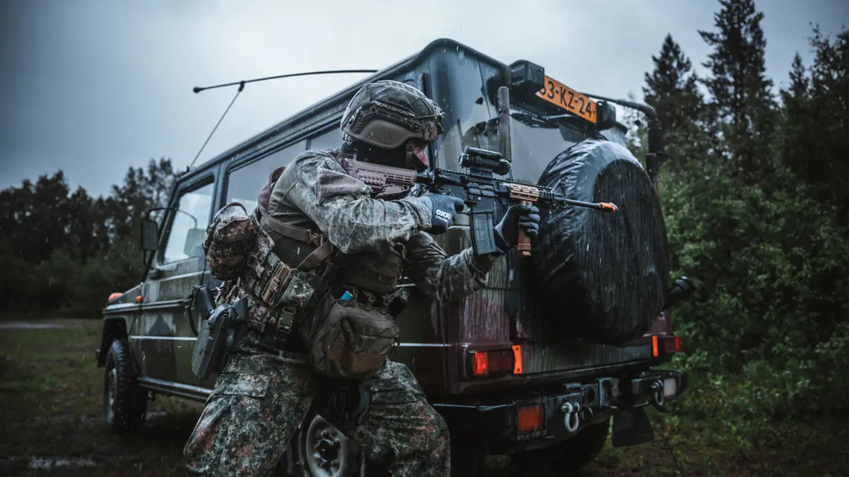 Een gewapende militair met een Colt voor zijn gezicht beweegt langs de achterkant van een groene terreinwagen.