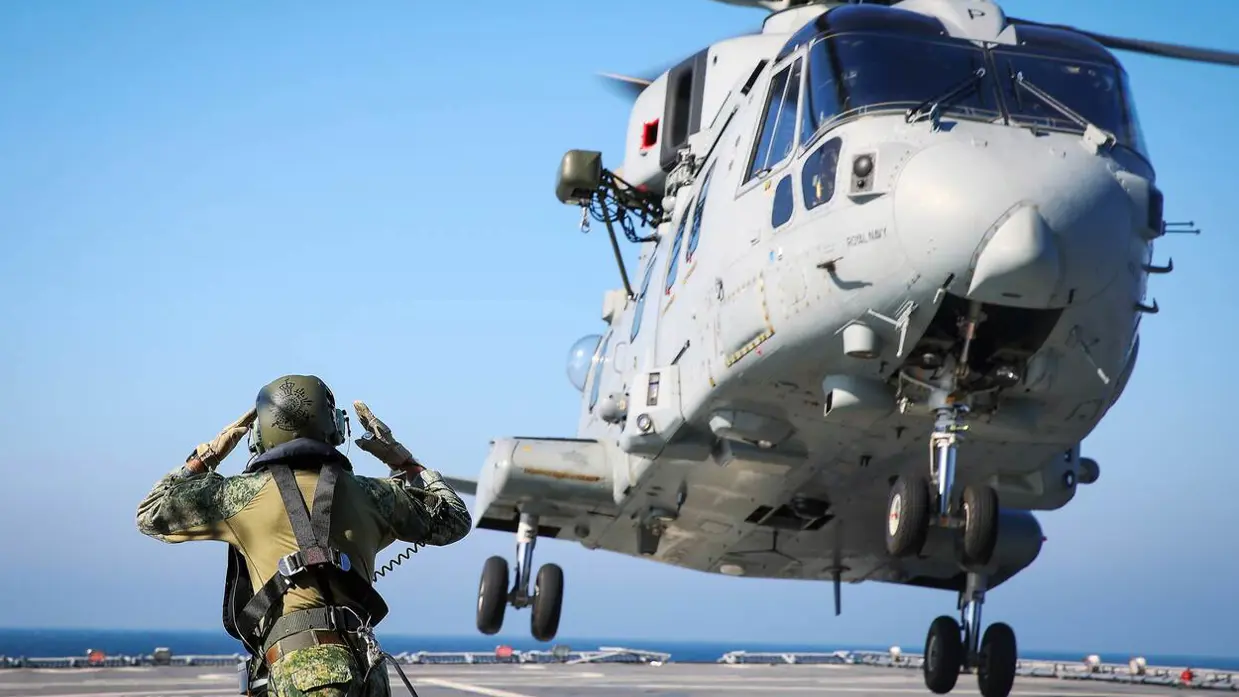Een lichtgrijze helikopter stijgt op, terwijl een militair op de voorgrond aanwijzingen geeft.