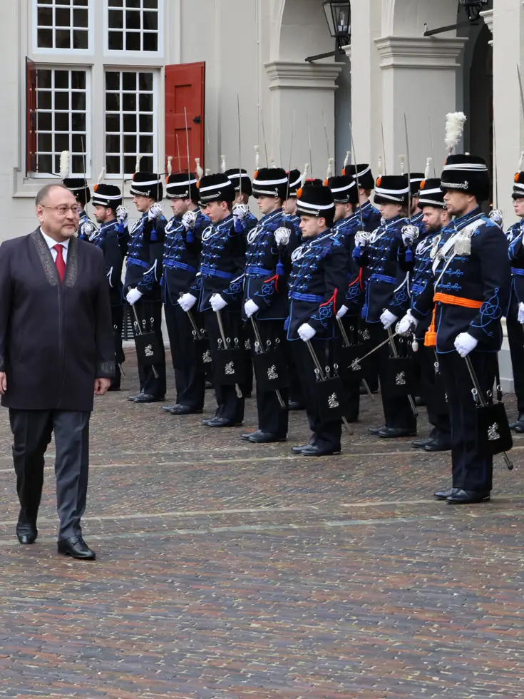 links: Een vrouw in een prachtige, versierde mantel loopt over een rode loper, terwijl een groep geüniformeerde wachten in blauwe tenues met sabel en rode pluim haar salueren. Midden: Een man in een blauwe, traditioneel gewaad loopt langs een rij wachten in blauwe uniformen met sabels en pluimen, voor een wit gebouw met rode luiken en een rode loper. Rechts: Een man in een donkere jas loopt langs een rij wachten in blauwe uniformen met pluimen en sabels, voor een wit gebouw met rode luiken en ramen.