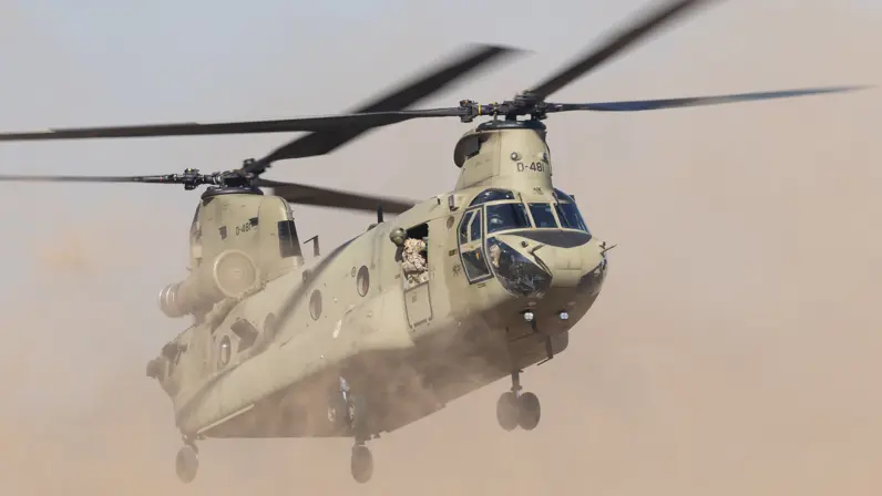 Een militaire helikopter vliegt laag boven een stoffige grond, met zand en stof die opwaaien door de rotorbladen.