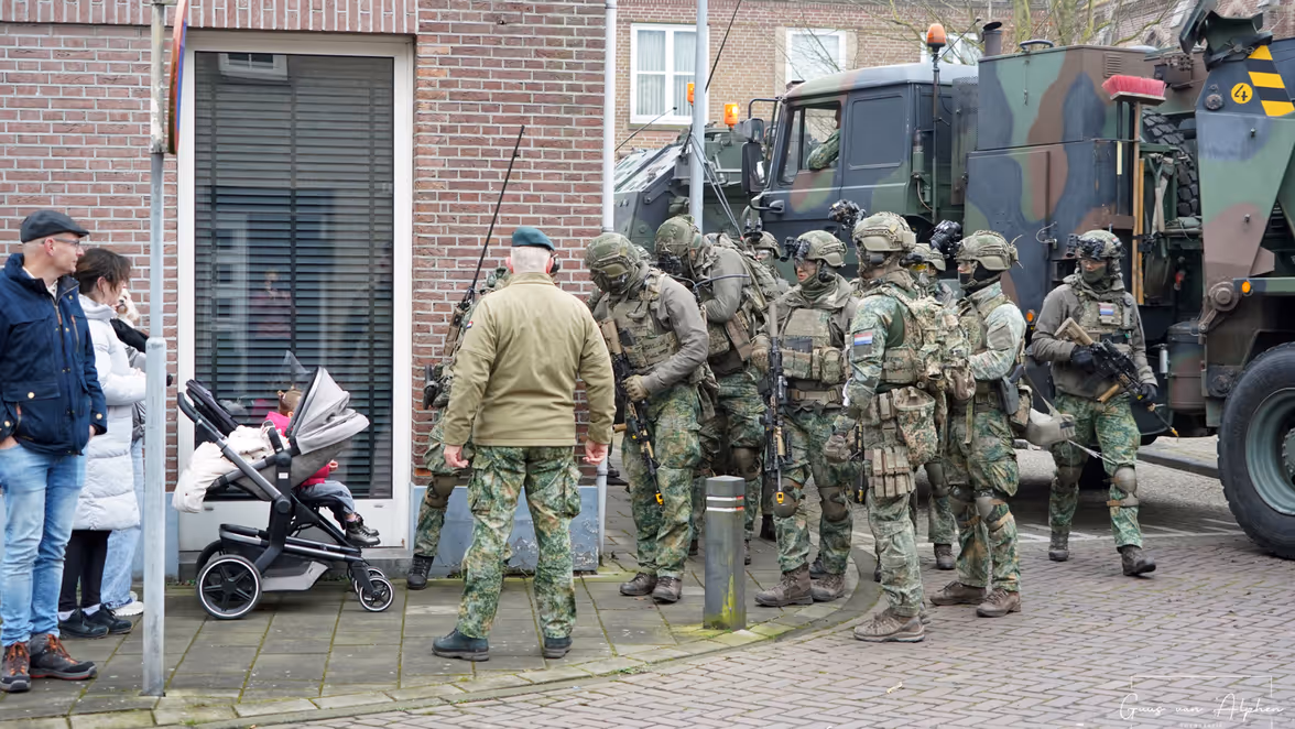 Zeven militairen met wapens staan naast een vrachtwagen en benaderen twee burgers in een gewone Nederlandse straat.