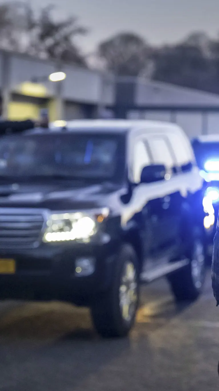 Een marechaussee staat bij een colonne dienstvoertuigen met ingeschakelde zwaailichten. 