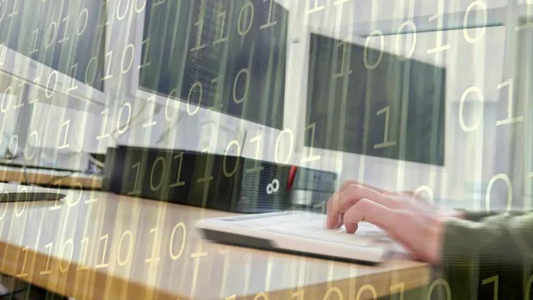 Een paar handen op een toetsenbord, achter computerschermen.