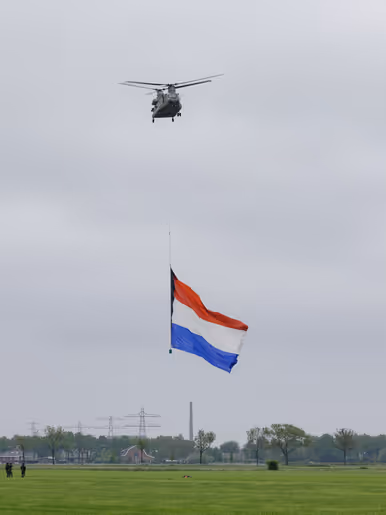 Een Chinook stijgt op boven een veld, met aan een lange kabel de Nederlandse vlag