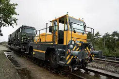 Een gele locomotief trekt groene Defensievrachtwagens.